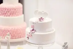 Biały tort dwupiętrowy z sercami i kwiatami, obok różowy tort.