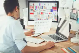 Mężczyzna, jako ux designer, analizuje wydruki przy biurku z komputerem i laptopem.