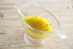 Vinagreta de mostaza dorada con hierbas verdes picadas, servida en una salsera de cristal sobre una mesa de madera.