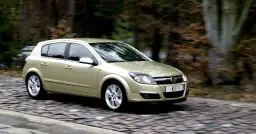 Złoty Opel Astra H 1.6 benzyna w ruchu na brukowanej drodze, otoczony jesiennym lasem.