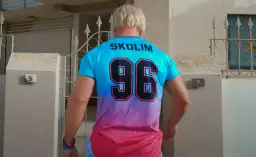 Mężczyzna w koszulce z napisem SKOLIM i numerem 96, stojący przed domem z numerem 5.