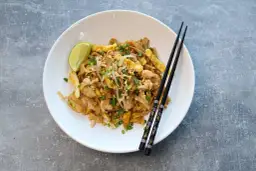 Pad thai z kurczakiem, jajkiem, orzeszkami i limonką na białym talerzu z pałeczkami.