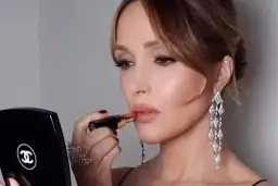 Elegancki makijaż na wesele: kobieta z błyszczącymi kolczykami i pierścionkiem nakłada szminkę, patrząc w lusterko Chanel.