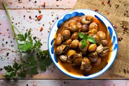 Un plato de caracoles en salsa, adornado con menta fresca, rodeado de especias y perejil sobre una mesa de madera.