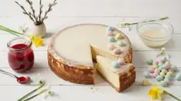 Pyszny sernik na Wielkanoc, udekorowany pastelowymi jajeczkami i lukrem. Obok słoiczek z konfiturą malinową i wiosenne kwiaty.