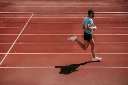Mężczyzna w niebieskiej koszulce i czarnych spodenkach biegnie po bieżni lekkoatletycznej.