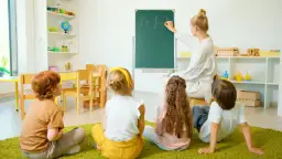 Nauczyciel wychowania przedszkolnego pisze na tablicy "1+1=2". Dzieci siedzą na dywanie i uważnie słuchają.