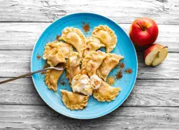 Pyszne pierogi z jabłkami, posypane cynamonem, na niebieskim talerzu, obok jabłka.