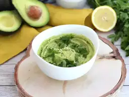 Un cremoso hummus de aguacate, decorado con perejil fresco y semillas de sésamo, junto a aguacates cortados y un limón.
