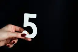 Dłoń z ciemnoczerwonym lakierem trzyma białą cyfrę 5, symbolizującą numerologię 5.