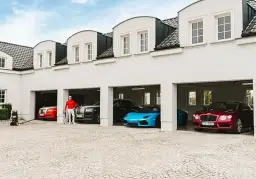 Piotr Śledź w czerwonej bluzie i białych spodniach stoi przy luksusowych samochodach: czerwonym Rolls-Royce, czarnym Rolls-Royce, niebieskim Lamborghini i czerwonym Bentleyu.