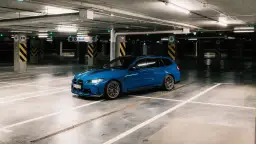 Niebieskie BMW M3 Touring z czarnymi pasami na masce stoi na pustym parkingu podziemnym.