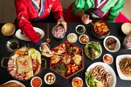 Dwie osoby w tradycyjnych koreańskich strojach jedzą obfity posiłek z wieloma daniami, w tym kimchi.
