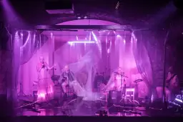 Na scenie teatru ludowego, w fioletowym świetle, artyści prezentują swój repertuar. Wokalistka w zwiewnej sukni i muzycy tworzą magiczną atmosferę.