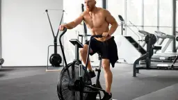Mężczyzna ćwiczy na rowerze stacjonarnym typu air bike w siłowni.
