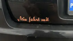 Pomarańczowa naklejka na czarnym lakierze samochodu z napisem "Mia fährt mit". Chcesz wiedzieć, jak usunąć naklejki z samochodu?