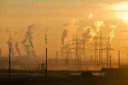 Dymiące kominy fabryk i linie energetyczne na tle zachodzącego słońca. To obraz przemysłowego krajobrazu, który ilustruje przyczyny zmian klimatu.