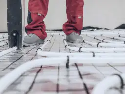 Pracownik w czerwonych spodniach i szarych butach stoi na siatce z rurkami ogrzewania podłogowego. Rozpoczyna się wygrzewanie posadzki po roku.