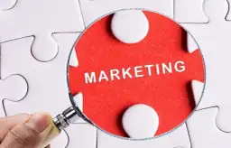 Pod lupą widać słowo "MARKETING" na czerwonym tle, otoczone białymi elementami układanki. To klucz do zrozumienia definicji marketingu.