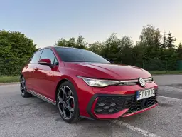 Czerwony Volkswagen Golf GTI na parkingu, z zielenią w tle.
