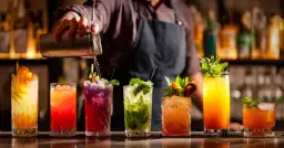Mistrz miksologii przygotowuje kolorowe drinki. Na blacie baru stoją drinki z owocami i miętą, a barman nalewa płyn z shakera.