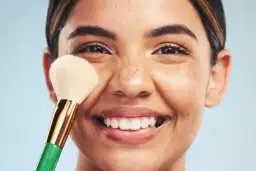 Una mujer sonríe mientras se aplica maquillaje con una brocha. Descubre trucos de maquillaje profesional para realzar tu belleza natural.