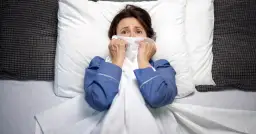 Kobieta w piżamie leży w łóżku, przerażona, z chusteczką przy ustach. Wygląda, jakby doświadczyła omdlenia we śnie.