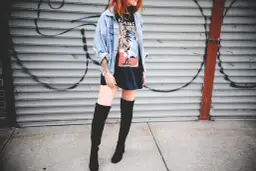 Chica con botas altas para bajitas, chaqueta vaquera y camiseta gráfica, posando frente a una pared de garaje con grafitis.