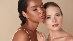 Dos mujeres, una con piel oscura y ojos cerrados, la otra con piel clara y ojos abiertos, comparten un momento íntimo sobre un fondo beige.