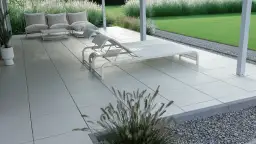Nowoczesny taras betonowy z leżakami i sofą, otoczony trawą i kamieniami. Idealne miejsce na relaks.