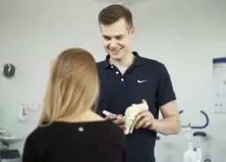 Uśmiechnięty technik fizjoterapii pokazuje pacjentce model stawu kolanowego, wyjaśniając szczegóły.