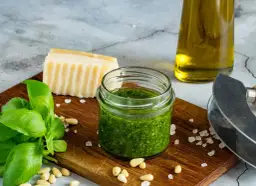 Słoiczek domowego pesto z bazylii, idealny sposób, jak zrobić pesto z bazylii na zimę. Obok świeża bazylia, orzeszki piniowe, parmezan i oliwa.