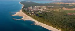 Jarosławiec atrakcje: aerial view of a coastal town with sandy beaches, breakwaters, pine forests, and a lake in the distance.