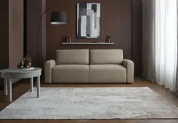 Elegancka, beżowa kanapa na tle brązowej, panelowej ściany w salonie. Nad nią abstrakcyjny obraz i półka z dekoracjami.