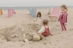 Dzieci budują zamek z piasku, ciesząc się beztroskimi zabawami na plaży. W tle widać leżaki i parawany.