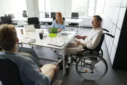 Zespół omawia projekty. Mężczyzna na wózku inwalidzkim i osoba z tabliczką Braille'a, pokazując, że **2 grupa inwalidzka przywileje w pracy** są uwzględniane.