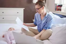 Kobieta w okularach pracuje na laptopie, podczas gdy dziecko śpi obok. Zastanawia się, czy można zameldować dziecko bez rodziców.
