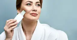 Kobieta w białym szlafroku używa urządzenia do peelingu kawitacyjnego. Dowiedz się, jak używać peeling kawitacyjny dla promiennej cery.