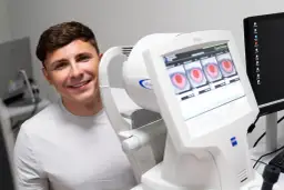 Uśmiechnięty mężczyzna obok urządzenia okulistycznego z obrazami oczu na ekranie.