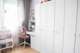 Urocza aranżacja małego pokoju z dużą białą szafą, biurkiem, różową kuchnią dla dzieci i plecakiem w kształcie sowy.