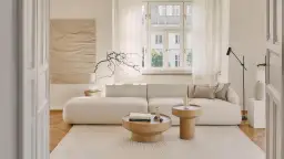 Jasny, przytulny salon z beżowym domem w tle. Miękka sofa, drewniane stoliki i subtelne dekoracje tworzą spokojną atmosferę.