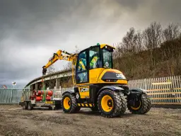 Żółta koparka JCB Hydradig, ważąca ok. 11 ton, ładuje rury na przyczepę.