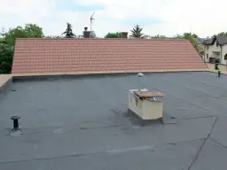 Płaski dach z papy, komin i fragment dachu skośnego z dachówką. To właśnie attyka co to za element?