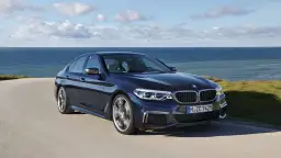 Ciemnoniebieskie BMW M550i sunie po krętej drodze nad brzegiem morza.