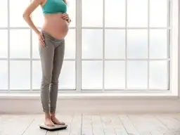 Kobieta w ciąży na wadze, zastanawia się, ile można przytyć w ciąży. Jasne światło wpada przez okno.