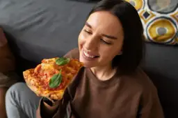 Uśmiechnięta kobieta delektuje się kawałkiem pizzy, jakby właśnie zakończyła jej odgrzewanie w piekarniku.