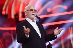 Jan Pietrzak w garniturze i okularach przeciwsłonecznych przemawia do mikrofonu na scenie.