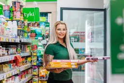 Kobieta w zielonej koszulce trzyma stos pudełek z pizzą na wynos w sklepie osiedlowym.