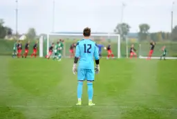 Golkiper w błękitnym stroju z numerem 12 czeka na akcję na boisku. W tle inni zawodnicy i bramka.