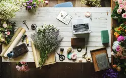 Kwiaty, laptop i akcesoria florystyczne, jak nożyczki, wstążki i sznurek, tworzą inspirujący warsztat. Widać też materiały do pakowania, przypominające **faszynę**.